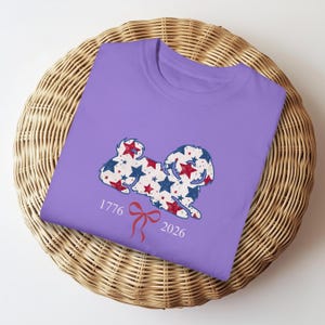 May include: A lilac t-shirt with a dog silhouette design filled with red, white, and blue stars. The design includes the numbers "1776" and "2026" with a red bow. The shirt is displayed on a woven basket.