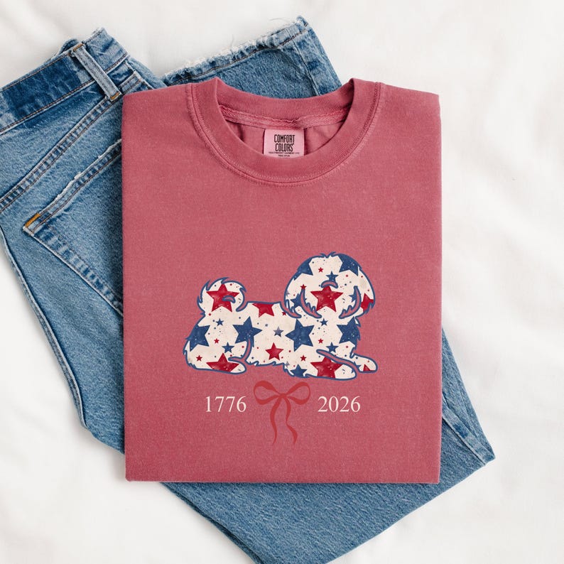 May include: A maroon t-shirt featuring a dog design adorned with red and blue stars. The numbers "1776" and "2026" are printed below the dog, with a red bow in the middle. The t-shirt is folded on top of a pair of blue denim jeans.