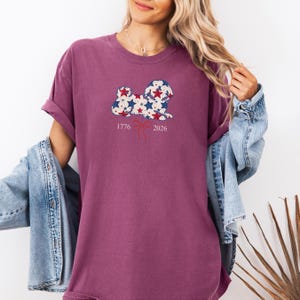 May include: A maroon t-shirt with a dog-shaped graphic filled with red, white, and blue stars. The numbers "1776" and "2026" are printed below the graphic, with a red ribbon design in between. The shirt has rolled-up sleeves.