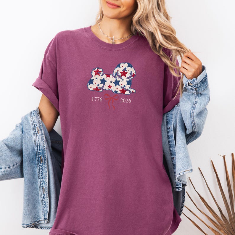 May include: A maroon t-shirt with a dog-shaped graphic filled with red, white, and blue stars. The numbers "1776" and "2026" are printed below the graphic, with a red ribbon design in between. The shirt has rolled-up sleeves.