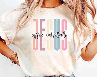 May include: A cream-coloured t-shirt featuring the word "JESUS" in pastel shades, with "coffee and pitbulls" written in a handwritten style. The shirt has a relaxed fit and short sleeves.
