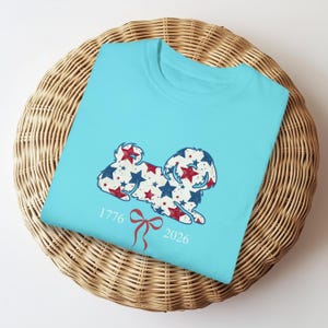 May include: A light blue t-shirt with a dog design made of red and blue stars. The shirt has the numbers "1776" and "2026" with a red bow graphic. The t-shirt is folded on a woven basket.