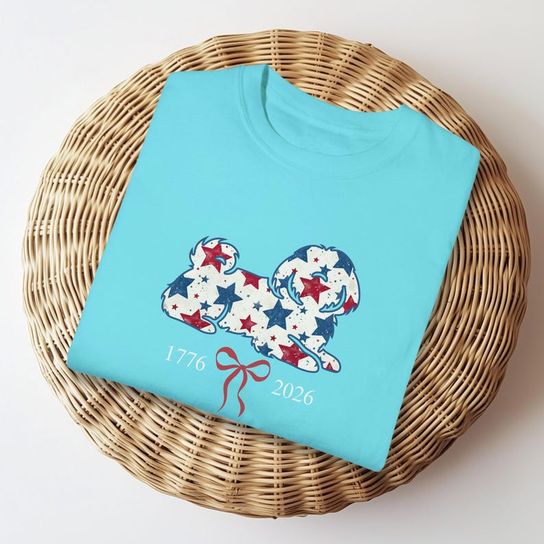 May include: A light blue t-shirt with a dog design made of red and blue stars. The shirt has the numbers "1776" and "2026" with a red bow graphic. The t-shirt is folded on a woven basket.