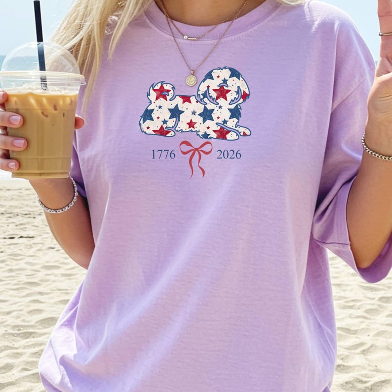 May include: A lilac t-shirt with a dog illustration composed of red and blue stars. The numbers "1776" and "2026" are printed below the dog, separated by a red bow. The shirt has a loose fit.