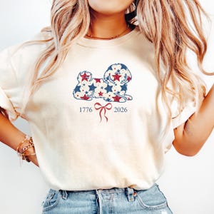 May include: A cream-coloured sweatshirt with a dog silhouette design filled with red and blue stars. The numbers "1776" and "2026" are below the dog, with a red bow. The top has long sleeves and a loose fit.