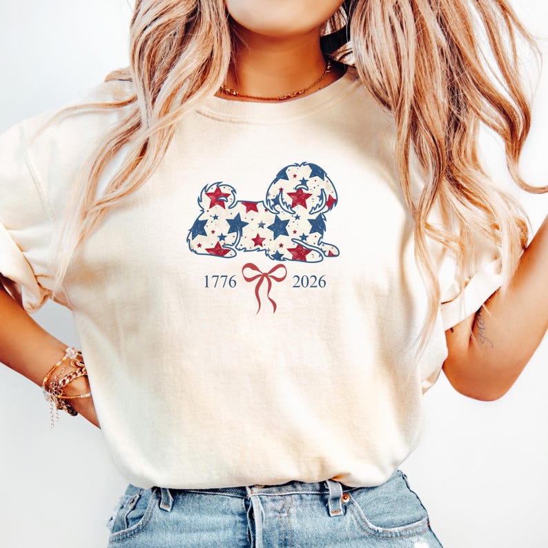 May include: A cream-coloured sweatshirt with a dog silhouette design filled with red and blue stars. The numbers "1776" and "2026" are below the dog, with a red bow. The top has long sleeves and a loose fit.