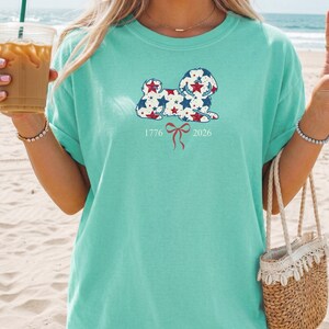 May include: A light teal t-shirt with a dog illustration decorated with red, white, and blue stars. The design includes the dates 1776 and 2026, and a red bow. The shirt is a casual style.