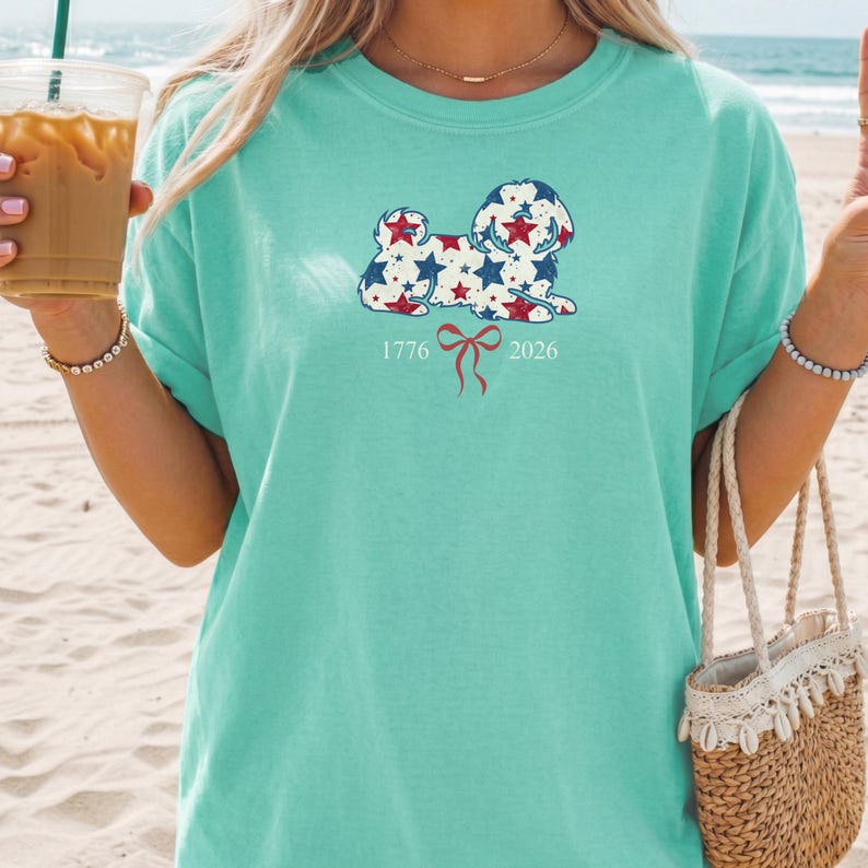 May include: A light teal t-shirt with a dog illustration decorated with red, white, and blue stars. The design includes the dates 1776 and 2026, and a red bow. The shirt is a casual style.