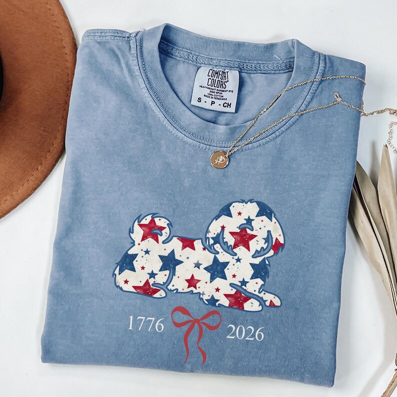 May include: A pale blue Comfort Colors t-shirt with a dog design made of red and blue stars. The shirt has the numbers "1776" and "2026" with a red bow. A gold necklace is draped across the shirt.
