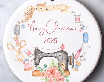 May include: White ceramic Christmas ornament with a sewing theme. The ornament features a watercolor design of a sewing machine, spools of thread, scissors, and the text "Merry Christmas 2025". A floral arrangement and measuring tape complete the design.