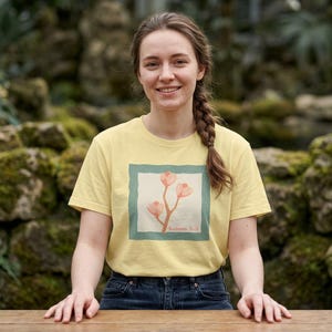 Puede incluir: Una camiseta de manga corta de color amarillo pálido con un gráfico cuadrado en el pecho. El gráfico muestra una ilustración estilizada de tres flores rosas sobre un tallo marrón con fondo claro, enmarcado por un borde verde azulado con el texto Salmo 5:3.