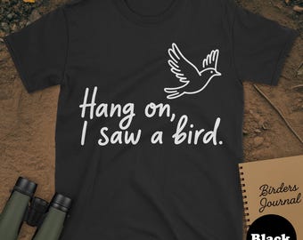 May include: Black t-shirt featuring the text "Hang on, I saw a bird" and a white bird illustration. A notebook with "Birders Journal" and binoculars are also in the image.