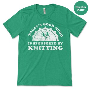Op de afbeelding: Groen T-shirt met een witte, verweerde afbeelding met de tekst "Today's Good Mood Is Sponsored By Knitting" met een cartoon zon gezicht.