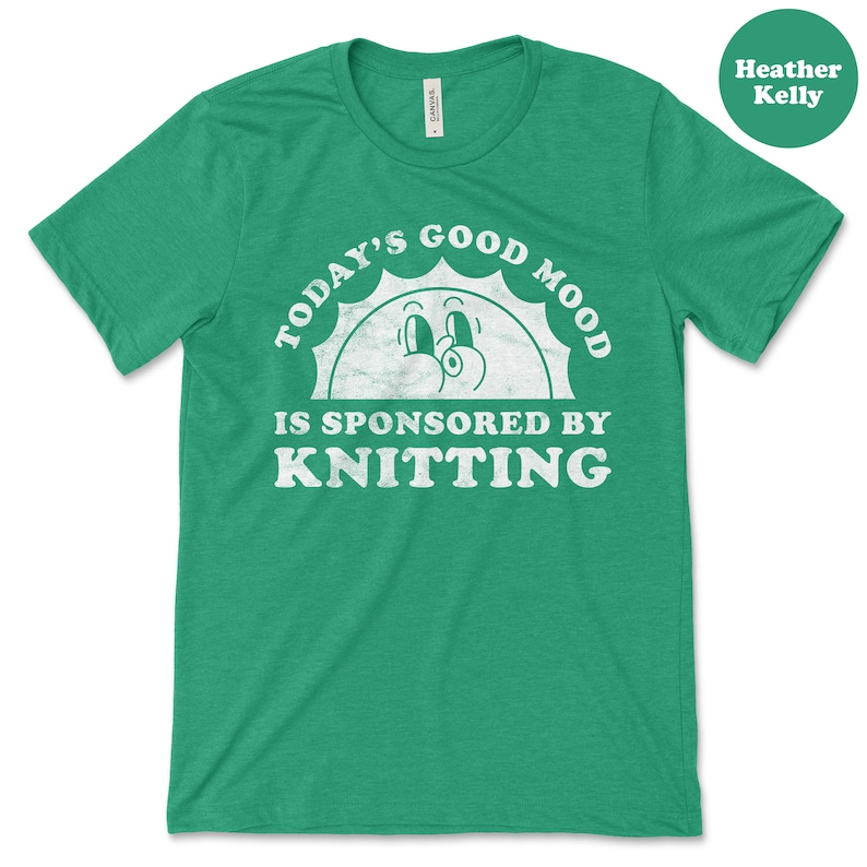 Op de afbeelding: Groen T-shirt met een witte, verweerde afbeelding met de tekst "Today's Good Mood Is Sponsored By Knitting" met een cartoon zon gezicht.