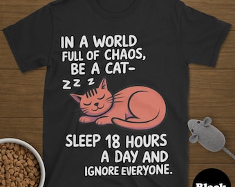 Puede incluir: Camiseta negra con un gato naranja durmiendo y el texto "IN A WORLD FULL OF CHAOS, BE A CAT - SLEEP 18 HOURS A DAY AND IGNORE EVERYONE." También se ve un ratón de juguete y un cuenco de comida para gatos.
