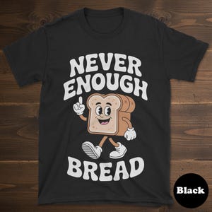 May include: Black t-shirt with the text "NEVER ENOUGH BREAD" in white, featuring a cartoon bread slice character. The bread slice has a smiling face, arms, and legs. The shirt has a round neck.