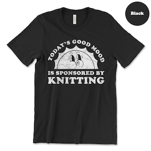 Op de afbeelding: Zwart T-shirt met een witte afbeelding waarop staat "Today's good mood is sponsored by knitting" met een cartoon zon gezicht.