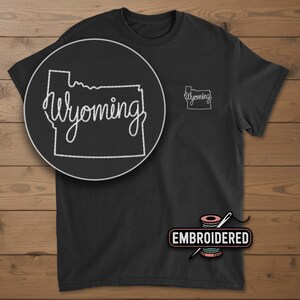 May include: Black t-shirt with a white embroidered outline of the state of Wyoming and the word "Wyoming" in cursive. A larger, circular close-up of the embroidery is on the left. The word "Embroidered" is below the shirt.