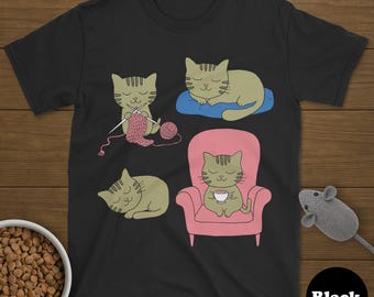 May include: Black t-shirt with a cartoon cat design. Four cats are depicted: one knitting, one resting on a blue cushion, one sleeping, and one enjoying a drink in a pink armchair. A toy mouse and a bowl of cat food are also visible in the image.