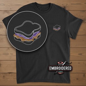 May include: Black t-shirt with an embroidered peanut butter and jelly sandwich design. The design is in a circle and also appears as a smaller version on the shirt. The word "EMBROIDERED" is printed on the shirt.