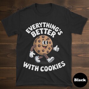 May include: Black t-shirt featuring the phrase "EVERYTHING'S BETTER WITH COOKIES" and a cartoon cookie character. The cookie has a smiling face, arms, legs, and is giving a thumbs up. The shirt is a crew neck.