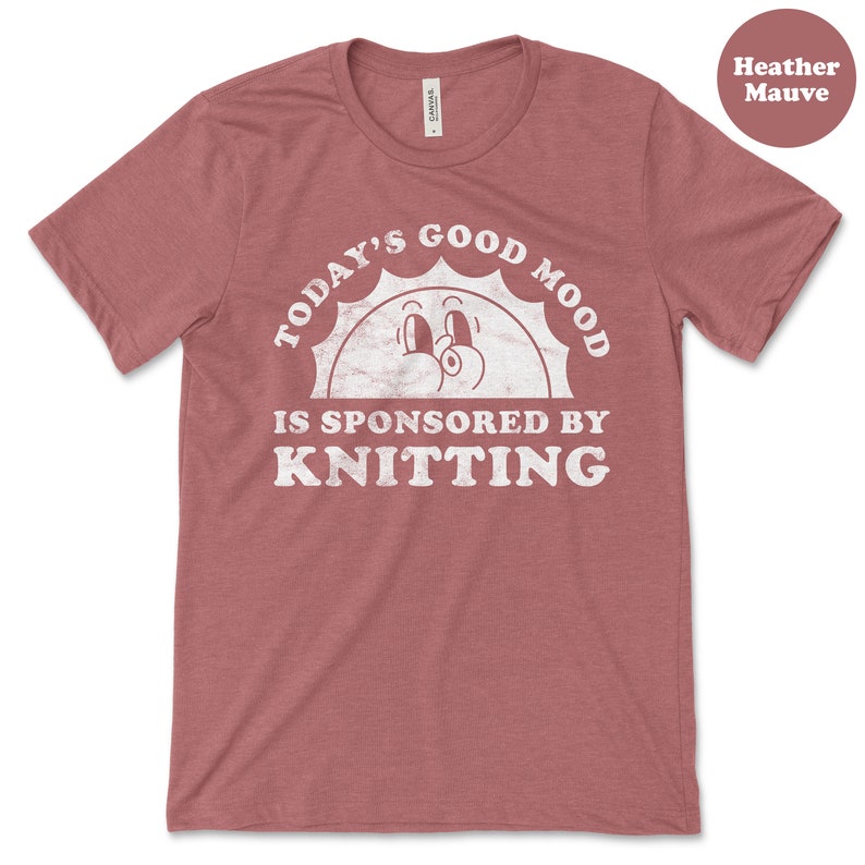 Op de afbeelding: Heather mauve t-shirt met een witte afbeelding met de tekst "Today's good mood is sponsored by knitting" met een cartoon zon met ogen en een glimlach.