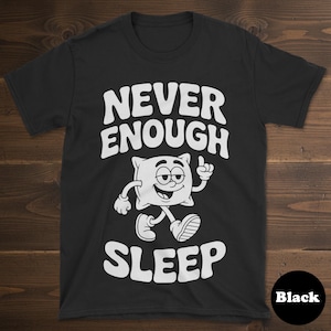 May include: Black t-shirt with the text "NEVER ENOUGH SLEEP" in white, above a cartoon pillow character. The pillow is white with a smiling face and is walking with one finger pointing up. The word "Black" is in a black circle.
