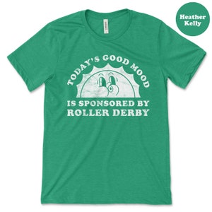 Op de afbeelding: Een groen T-shirt met een witte afbeelding waarop staat "Today's Good Mood Is Sponsored By Roller Derby". De afbeelding toont een cartoonzon met ogen en een glimlach.
