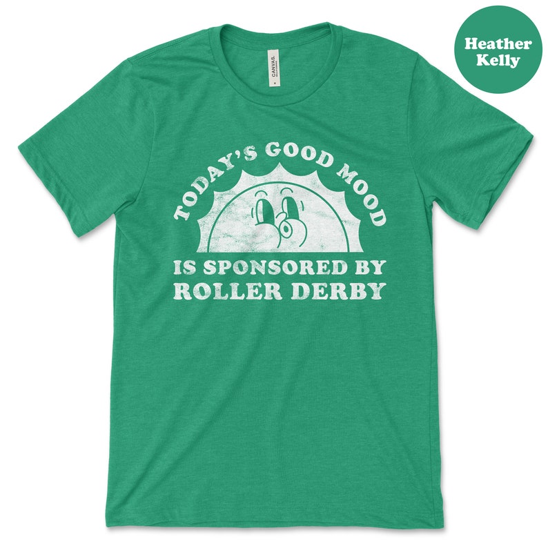 Op de afbeelding: Een groen T-shirt met een witte afbeelding waarop staat "Today's Good Mood Is Sponsored By Roller Derby". De afbeelding toont een cartoonzon met ogen en een glimlach.