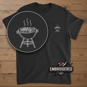 May include: Black t-shirt with a white embroidered design of a barbecue grill with sausages and steam. The shirt has a small grill design on the chest and the word "EMBROIDERED" below the design.