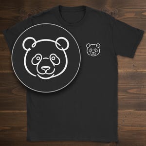 May include: A black short-sleeved t-shirt featuring a white line art graphic of a panda bear face on the left chest. The design is rendered in a single continuous line style, showcasing the panda's ears, eyes, nose, and mouth clearly.