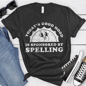May include: A dark gray t-shirt with a white graphic of a smiling sun with the text "Today's Good Mood Is Sponsored By Spelling".