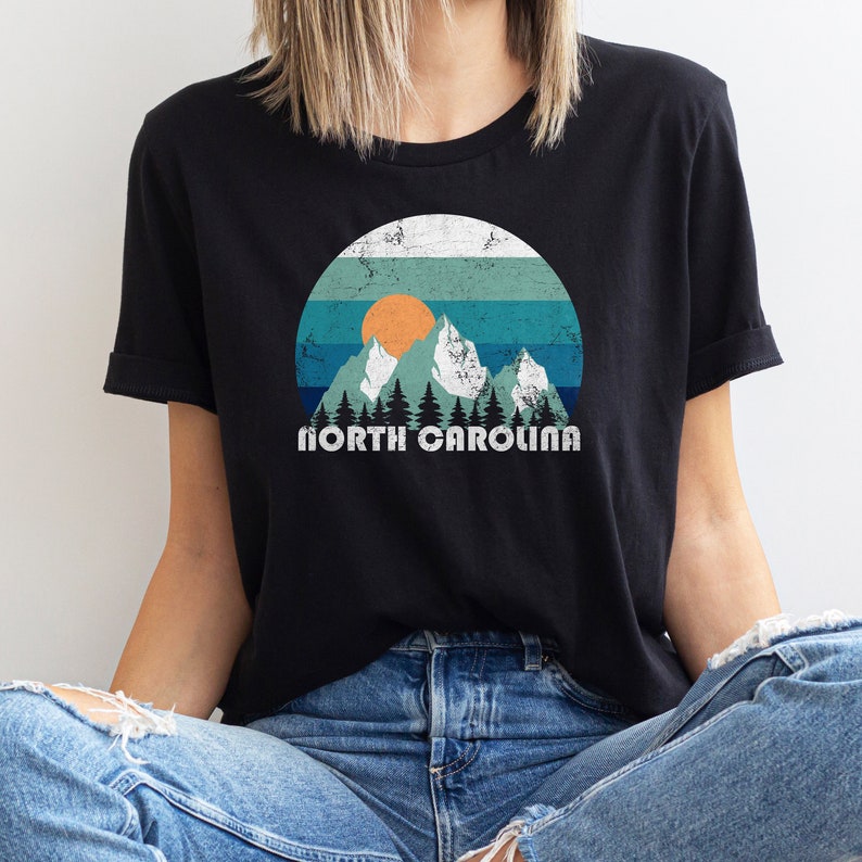 May include: A black t-shirt with a vintage-style graphic of mountains, a sun, and trees. The text "NORTH CAROLINA" is printed below the graphic.