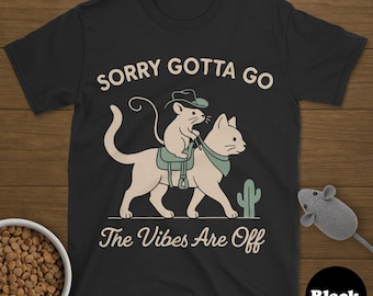May include: Black t-shirt with a humorous graphic of a mouse riding a cat, wearing a cowboy hat and saddle. The text reads "Sorry Gotta Go, The Vibes Are Off". A cactus is also in the graphic.