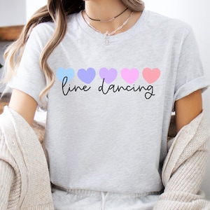 May include: Light gray t-shirt with the words "line dancing" in a cursive font. Above the text are five pastel-colored hearts in blue, purple, pink, and peach. The shirt has short sleeves and a crew neck.