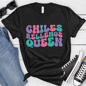 May include: Black t-shirt with the text "Chiles Rellenos Queen" in a retro font with pink, purple, and teal colors.