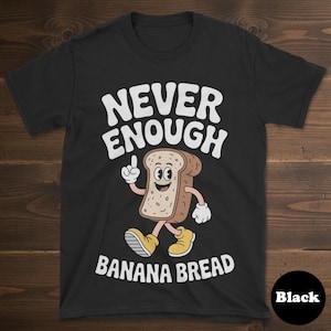 May include: Black t-shirt with the text "NEVER ENOUGH BANANA BREAD" in white. The shirt features a cartoon slice of bread with a smiling face, arms, and legs. The bread is wearing yellow shoes and pointing upwards.