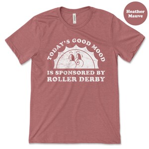 Op de afbeelding: Heather mauve t-shirt met een witte afbeelding van een lachende zon met de tekst "Today's Good Mood Is Sponsored By Roller Derby".