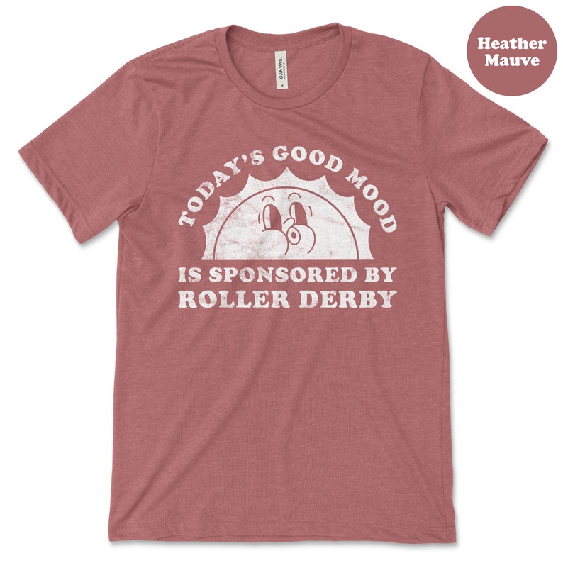 Op de afbeelding: Heather mauve t-shirt met een witte afbeelding van een lachende zon met de tekst "Today's Good Mood Is Sponsored By Roller Derby".