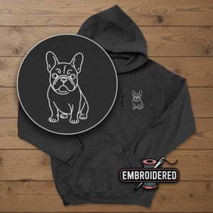 May include: A dark gray hoodie with a white embroidered French Bulldog design on the chest and a larger circular design. The hoodie has a front pocket and a hood. The word "EMBROIDERED" is printed on a label.
