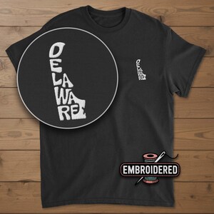 May include: Black t-shirt with a white embroidered outline of the state of Delaware. The word "DELAWARE" is vertically embroidered within the state's shape. A smaller version of the design is on the chest. The word "EMBROIDERED" is below the shirt.