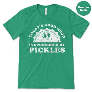 May include: Green t-shirt with a white graphic that says "Today's Good Mood Is Sponsored By Pickles". The graphic features a cartoon sun with a smiling face.