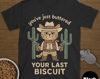 Peut inclure: T-shirt noir avec un chat de dessin animé vêtu en cow-boy, tenant deux pistolets, avec des cactus. Le texte dit "you've just buttered YOUR LAST BISCUIT". Un bol de nourriture pour chat et une souris jouet sont également dans l'image.