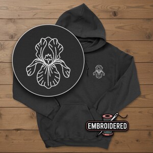 May include: A black hoodie with a white embroidered iris flower design on the chest and a larger magnified view in a circle. The word "EMBROIDERED" is on a label near the bottom.