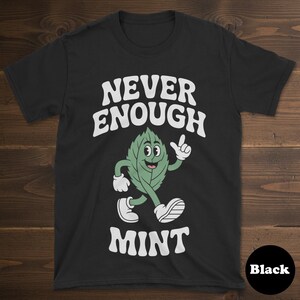 May include: Black t-shirt featuring the text "NEVER ENOUGH MINT" in white, with a cartoon mint leaf character. The mint leaf has a smiling face, white gloves, and shoes. The word "Black" is in a black circle.