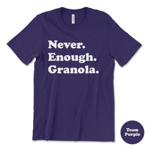 May include: A purple t-shirt with white text that reads "Never. Enough. Granola." The shirt is a crew neck style with short sleeves.