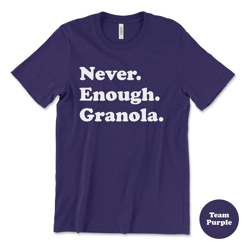 May include: A purple t-shirt with white text that reads "Never. Enough. Granola." The shirt is a crew neck style with short sleeves.