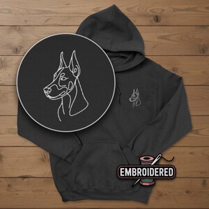May include: A dark gray hoodie with a white embroidered outline of a Doberman Pinscher dog on the chest and a larger version in a circular patch. The word "EMBROIDERED" is on a label at the bottom.