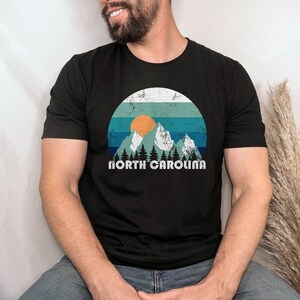 May include: A black t-shirt with a vintage-style graphic of mountains, a sunset, and trees. The text "NORTH CAROLINA" is printed below the graphic.
