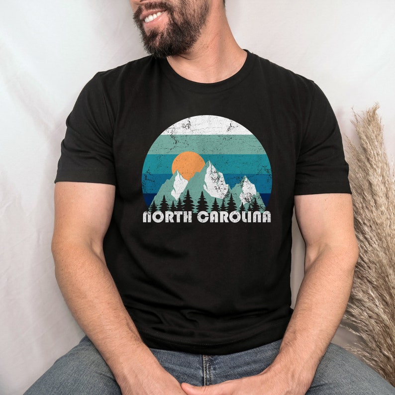 May include: A black t-shirt with a vintage-style graphic of mountains, a sunset, and trees. The text "NORTH CAROLINA" is printed below the graphic.